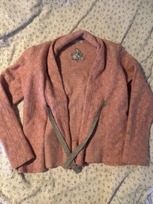 Rosa kofta från Odd Molly - Mysig rosa kofta från Odd Molly med grå knytband framtill och broderad logga i nacken. Koftan har en avslappnad passform, långa ärmar och mjukt material som känns skönt mot huden. Perfekt för lager på lager och en chill look. Storlek S/M