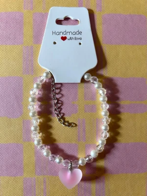 Pärlarmband med rosa hjärthänge - Sött armband med vita pärlor och transparenta glaspärlor, dekorerat med ett rosa hjärthänge. Armbandet har justerbar kedja i silverfärg och är handgjort. Perfekt accessoar för en trendig look.
