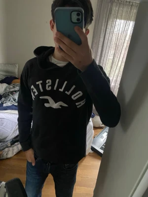 Svart Hollister hoodie med logga - Svart hoodie från Hollister med stor vit logga och fågeltryck på bröstet. Klassisk känguruficka framtill och huva med dragsko. Perfekt för en avslappnad och stilren look.