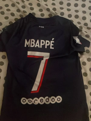 PSG Mbappé matchtröja Nike - Snygg PSG matchtröja med Mbappé och nummer 7 på ryggen. Tröjan är mörkblå med vita och röda detaljer, PSG-logga på bröstet och Qatar Airways som sponsor. Tillverkad i lätt och ventilerande material, perfekt för fotboll eller träning. Priset kan pratas 