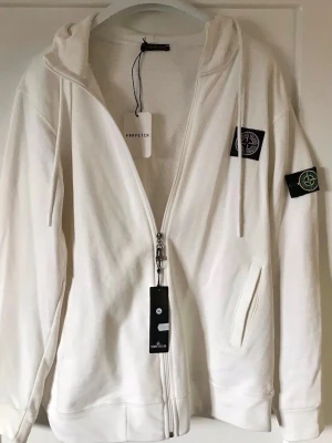 Vit hoodie från Stone Island - Snygg vit hoodie från Stone Island med dragkedja, huva och klassiska logopatchar på bröstet och ärmen. Tillverkad i mjukt bomullsmaterial och har två fickor framtill. Perfekt för en avslappnad och trendig stil.