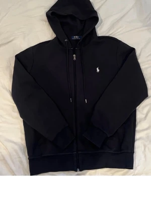Svart zip hoodie från Polo Ralph Lauren - Klassisk svart hoodie med dragkedja från Polo Ralph Lauren. Hoodien har huva med dragsko, två fickor framtill och den ikoniska vita Polo-loggan broderad på bröstet. Perfekt för en avslappnad och stilren look.