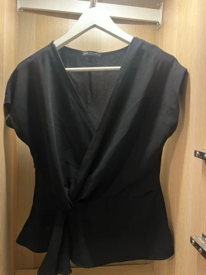 Svart omlottopp från Zara - Svart omlottopp från Zara med v-ringning. Toppen har korta ärmar och knyt i midjan för en snygg siluett. Perfekt att styla med jeans eller kjol för en trendig look. 