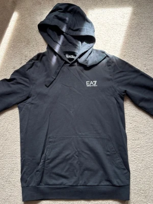 Svart hoodie från EA7 Emporio Armani - Svart hoodie från EA7 Emporio Armani med vit logga på bröstet och diskreta ränder baktill. Klassisk känguruficka framtill, dragsko i huvan och mjukt material. Perfekt för en chill och stilren look.