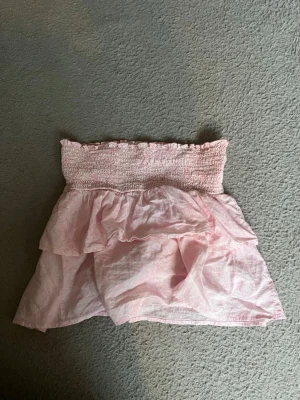 Ljusrosa volangkjol med smock - Använd gärna köp nu🩷 Söt ljusrosa kortkjol med smock i midjan och dubbla volanger. Kjolen har en luftig och lätt känsla, perfekt för varma dagar. Passar till både sneakers och sandaler för en avslappnad look.