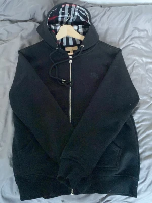 Svart hoodie från Burberry - Svart hoodie med dragkedja från Burberry. Klassisk huva med det ikoniska rutmönstret i svart, vitt och rött på insidan. Mjuk bomullskänsla, fickor framtill och justerbar dragsko i huvan. Perfekt för en stilren och avslappnad look.
