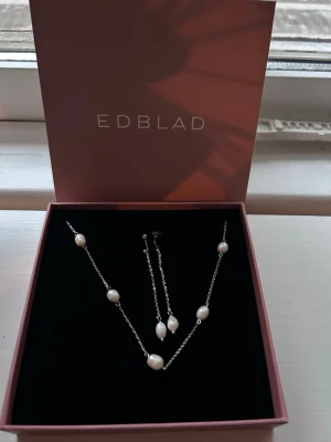 Smyckesset med pärlor från Edblad - Snyggt smyckesset från Edblad med halsband och örhängen i silverfärgad metall och vita pärlor. Kedjan är tunn och elegant, och pärlorna är ovala och jämnt placerade. Perfekt för dig som gillar stilrena accessoarer med en touch av klass. Örhängen är inte från Edblad men köpte i Åhléns äkta söttvattenspärlor och stål 