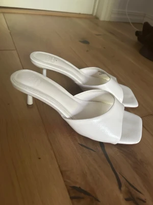 Vita slip-in pumps med smal klack - Säljer ett par trendiga vita slip-in pumps med öppen tå och fyrkantig framdel. Skorna har en smal klack och bred rem över foten. Tillverkade i ett glansigt material som ger en stilren look. Perfekta för dig som vill ha en clean och modern stil.