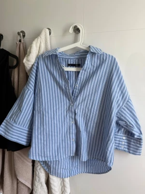Blårandig skjorta från 157 - Säljer en oversized skjorta från 157 i blått och vitt med klassiskt randigt mönster. Skjortan har knappar framtill, krage och trekvartsärmar med knappdetalj vid ärmslut. Perfekt för en avslappnad och trendig look.
