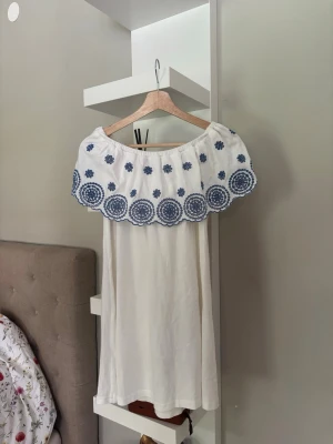 Vit offshoulder klänning med blå brodyr - Säljer en söt vit offshoulder klänning med bred volangkant dekorerad med blå broderade blommor och cirklar. Klänningen har en avslappnad passform och är perfekt för varma dagar. Materialet känns som bomull och ger en luftig känsla.
