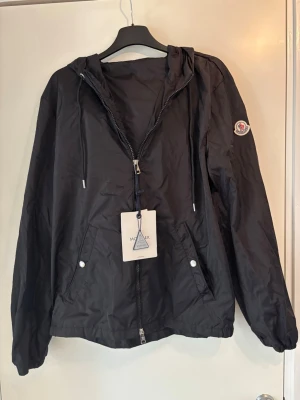 Svart vindjacka från Moncler - Svart vindjacka från Moncler med huva och dragkedja framtill. Jackan har två fickor med tryckknappar, justerbar dragsko i huvan och Moncler-logga på ärmen. På huvan finns en snygg detalj med röd och vit rand. Tillverkad i lätt, vattenavvisande material. Säljer pga för stor för mig. Pris kan diskuteras! 