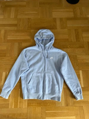 Ljusblå zip hoodie från Nike - Snygg ljusblå hoodie från Nike med dragkedja framtill och klassisk huva med dragsko. Har två stora fickor på magen och Nike-logga broderad i vitt på bröstet. Perfekt för en chill och sportig look. Tillverkad i mjukt bomullsmaterial.