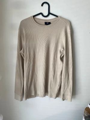 Beige långärmad tröja från H&M - En stilren beige långärmad tröja från H&M i ribbat material. Tröjan har rund halsringning och breda muddar vid ärmsluten. Perfekt för lager-på-lager och enkel att matcha med olika outfits.