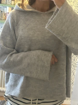Ljusgrå stickad hoodie - Mysig ljusgrå stickad hoodie med luva och vida ärmar. Tröjan har en avslappnad passform och är tillverkad i ett mjukt, fluffigt material som känns skönt mot huden. Perfekt för lager på lager och chill dagar.