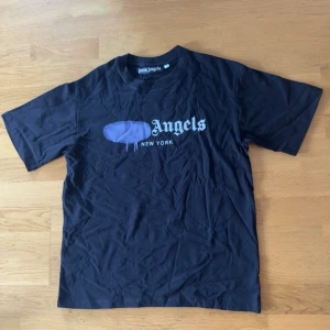 Svart Palm Angels t-shirt New York - Säljer en svart t-shirt från Palm Angels med trycket 'Angels New York' på bröstet och en lila grafisk detalj ovanför texten. T-shirten har klassisk passform och rund hals. Perfekt för dig som gillar streetwear och vill sticka ut.