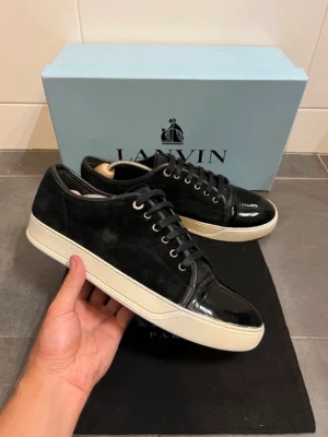 Svarta Lanvin sneakers i mocka och lack - Snygga svarta sneakers från Lanvin med ovandel i mocka och detaljer i lackat läder. Skorna har vita sulor och svarta skosnören. Perfekta för dig som gillar stilrena och exklusiva sneakers med en twist.
