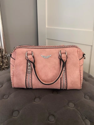 Rosa handväska - Snygg rosa handväska i fejkskinn med dubbla handtag och silverfärgade detaljer. Väskan har dekorativa nitar längs sidorna, svarta kontrastsömmar och ett emblem framtill. Perfekt storlek för det viktigaste och stängs med dragkedja.