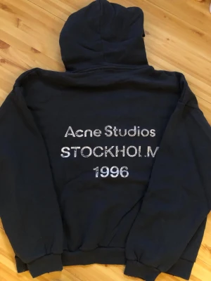 Svart hoodie Acne Studios Stockholm - Svart hoodie från Acne Studios med tryck på ryggen: 'Acne Studios STOCKHOLM 1996' i vitt. Klassisk modell med huva och muddar vid ärmslut och nederkant. Perfekt för en avslappnad och trendig look.