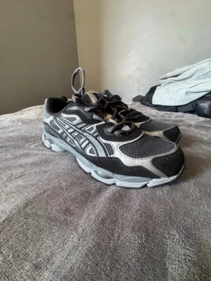 Svarta och grå sneakers från Asics - Snygga sneakers från Asics i svart och grå med klassiska ränder på sidan. Skorna har meshpaneler för bra ventilation och detaljer i syntetmaterial samt mocka. Perfekta för dig som gillar sportig stil och vill ha bekväma skor med snörning.