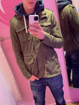 Olivgrön fieldjacka med fickor - Snygg olivgrön fieldjacka med fyra framfickor och dragkedja. Jackan har en klassisk rak passform och axelklaffar för extra stil. Perfekt för dig som vill ha en enkel men trendig look. 
