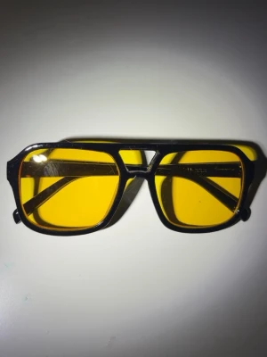 Svarta solglasögon med gula glas - Snygga solglasögon med svart båge och rektangulära gula glas. Glasögonen har en cool retrostil med markerad näsbrygga och breda skalmar. Perfekta för dig som vill sticka ut med en unik accessoar.