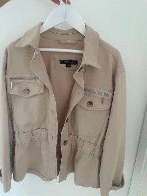 Beige jacka - Stilren beige overshirt från Comma med knappar framtill och två bröstfickor med både knapp och dragkedja. Jackan har en markerad midja med resår och klassisk krage. Perfekt för lager på lager och enkel att matcha💗
