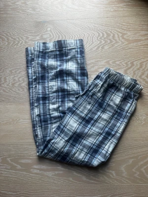 Rutiga pyjamasbyxor i blått och vitt - Mysiga pyjamasbyxor med klassiskt rutmönster i blått, vitt och grått. Byxorna har elastisk midja med dragsko och raka ben. Perfekta för chillkvällar hemma. Materialet känns mjukt och bekvämt mot huden. Har inte använts mycket 