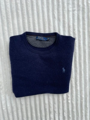 Mörkblå stickad tröja Polo Ralph Lauren - Mörkblå stickad tröja från Polo Ralph Lauren med rund halsringning och diskret broderad logga på bröstet. Klassisk och enkel design med ribbade muddar vid ärmslut och nederkant. Perfekt för lager på lager och stilrena outfits.
