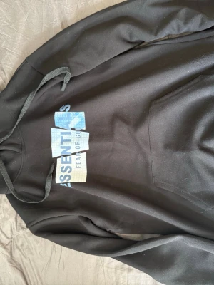 Svart Essentials Fear of God hoodie - Säljer en svart hoodie från Essentials Fear of God med stor logga i blått och vitt framtill. Hoodien har huva med dragsko och en klassisk magficka. Materialet känns mjukt och skönt, perfekt för en avslappnad streetwear-look.