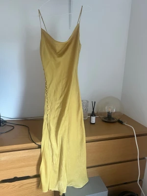 Gul långklänning med knappar - Snygg gul långklänning i glansigt material med smala axelband och dekorativa knappar längs sidan. Klänningen har en slits nedtill och en elegant, figurnära passform. Perfekt för sommarens alla tillfällen. Det saknas en knapp som man kan se på en bild💕