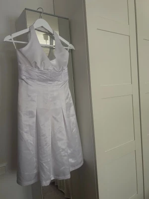 Vit halterneck klänning i satin - Stilren vit halterneck klänning i glansigt satinmaterial med v-ringning och markerad midja. Klänningen har en elegant siluett och dragkedja i ryggen. Perfekt för sommarens speciella tillfällen.