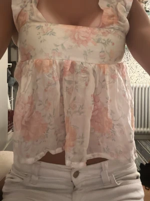 Blommig topp i pastell med rosett och öppen rygg - Söt hm topp med blommigt mönster i ljusa pastellfärger. Toppen har volangdetaljer vid axlarna, är ärmlös, rosett bak och öppen rygg och har en luftig passform. Perfekt för sommar och vår. Tillverkad i ett tunt bomullstyg.