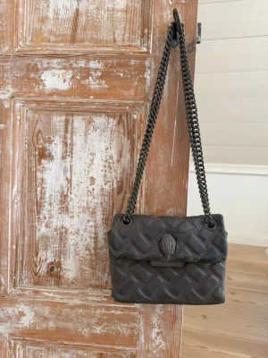 Grå Kurt Geiger väska - Snygg grå axelväska med quiltat mönster och grå kedjerem. Väskan har lock med dekorativt emblem och är tillverkad i ett mjukt, vadderat material. 
