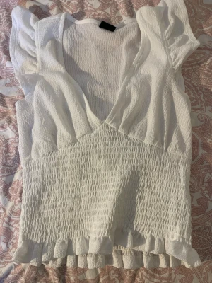 Vit smockad topp från Gina Tricot - Säljer en vit topp från Gina Tricot med smockad midja, volangkant nedtill och korta puffärmar. Djup v-ringning och mjukt, stretchigt tyg som ger en snygg passform. Perfekt för dig som gillar feminina detaljer och enkel stil.