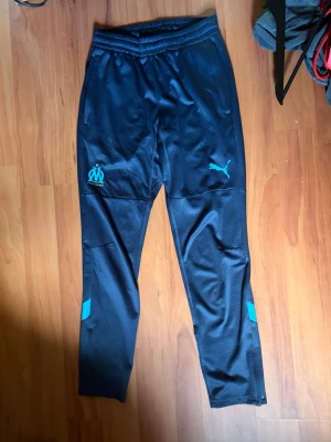 Svarta träningsbyxor Puma Marseille - Säljer ett par svarta träningsbyxor från Puma med Olympique de Marseille-logga i turkos på ena benet och Puma-logga på andra. Byxorna har elastisk midja, smal passform och detaljer i turkos vid benen. Perfekta för fotbollsträning eller gymmet.