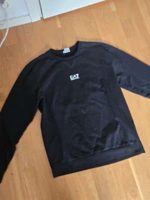 Svart sweatshirt från EA7 Emporio Armani - Snygg svart sweatshirt från EA7 Emporio Armani med vit logga på bröstet. Klassisk rund halsringning och långa ärmar. Perfekt för en stilren och avslappnad look. Materialet känns mjukt och bekvämt mot huden. Storlek L passarS/ M bättre