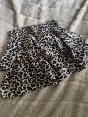 Kort leopardmönstrad kjol med volang - Säljer en kort kjol med leopardmönster i svart, beige och brunt. Kjolen har bred resår i midjan och volangdetaljer som ger en snygg och trendig look. Perfekt för dig som vill sticka ut lite extra.