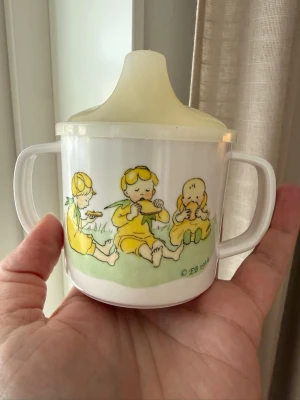 Vit pipmugg med blommotiv - Elsa Beskows motiv ”Blomsterflickor”, en  pipmugg  med två handtag och lock. Perfekt för småbarn som lär sig dricka själva. Motiven är klassiska och ger en nostalgisk känsla. Fint skick! Kanske något blekare än ny men ändå väldigt fint omhändertagen.