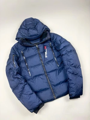 Polo Ralph Lauren RLX "Down Jacket" Jacka - Polo Ralph Lauren RLX Down Jacket | Modell: RLX Water-Resistant Down Jacket - Dunjacka med luva | Skick: Mycket fint skick / Sparsamt använd | Färg: Marinblå (Navy) med vita och röda detaljer | Material: Yttertyg 100% Nylon, Fyllning 75% Dun / 25% Fjäder | Nypris: ca 4 500 kr | Storlek: M - Passar även S | Hör av dig vid minsta fundering eller fråga 📦