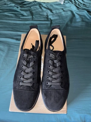 Christian Louboutin skor - Snygga svarta sneakers i mocka från Christian Louboutin med klassisk låg profil och ikonisk röd sula med signatur. Skorna har svarta skosnören och diskret branding på insidan. Perfekta för dig som vill ha en clean och exklusiv look. Köpta på vinted, pris går att diskutera vid snabb affär.
