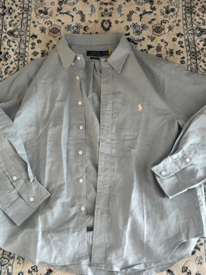 Ljusblå linneskjorta från Polo Ralph Lauren - Klassisk ljusblå skjorta från Polo Ralph Lauren med den ikoniska vita broderade loggan på bröstet. Skjortan har knappar framtill, lång ärm med manschett och är tillverkad i mjuk bomull. Perfekt för en clean och stilren look. Helt ny, äkta, tvättlapp är avklippt.