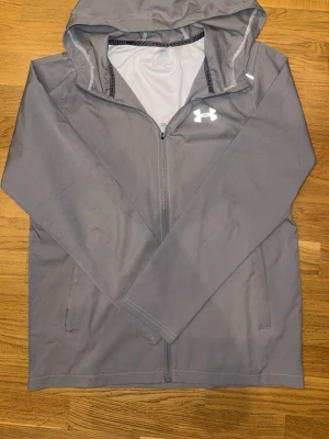 Grå vindjacka från Under Armour - Snygg grå vindjacka från Under Armour med huva och dragkedja framtill. Jackan har vita logotyper på bröstet och ryggen, samt två sidofickor. Tillverkad i lätt och funktionellt material som passar perfekt för aktiva dagar utomhus.
