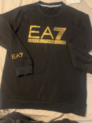 Svart EA7 Emporio Armani sweatshirt - Säljer en svart sweatshirt från EA7 Emporio Armani med guldfärgad logga på bröstet och ärmen. Tröjan har rund halsringning och ribbade muddar vid ärmslut och nederkant. Tillverkad i mjukt bomullsmaterial för en skön och avslappnad look.