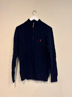 Mörkblå kabelstickad tröja Polo Ralph Lauren - Mörkblå kabelstickad tröja från Polo Ralph Lauren med halv dragkedja och röd broderad logga på bröstet. Tröjan har hög krage och ribbstickade muddar. Perfekt för en stilren och klassisk look.