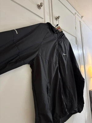 Svart vindjacka från Nike med huva - Snygg svart vindjacka från Nike med dragkedja framtill och huva. Jackan har en minimalistisk design med Nike-logga på bröstet och reflekterande detalj på ärmen. Tillverkad i lätt och vindtätt material, perfekt för blåsiga dagar.