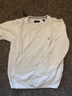 Vit stickad långtröja från GANT - Vit stickad tröja från GANT med rund halsringning och diskret logga broderad på bröstet. Tröjan har ribbade muddar vid ärmslut och nederkant samt en snygg detalj med mörkblå rand på insidan av kragen. Perfekt för en clean och klassisk stil.
