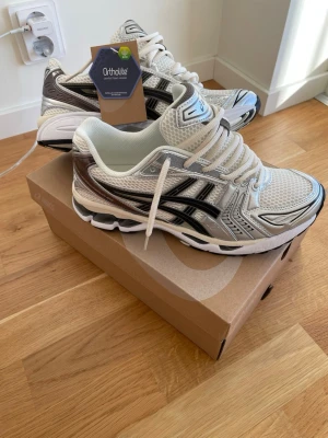 Asics GEL-Kayano 14  - Snygga Asics GEL-Kayano 14 sneakers i vitt och silver med svarta detaljer. Ovandel i mesh och syntet för bra andningsförmåga och komfort. Klassisk retrostil med Ortholite-innersula för extra skön känsla. Perfekta för dig som gillar sportig och trendig look.