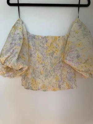 Blommig offshoulder topp från H&M - Supersöt offshoulder topp från H&M med puffiga ärmar och blommigt mönster i pastelliga toner av gul, lila och vit. Smockad kropp för en bekväm och figurnära passform. Perfekt för vår och sommar när du vill känna dig extra fin.