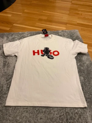 Vit HUGO t-shirt med tryck - Vit t-shirt från HUGO med rött logotryck och svart motiv på bröstet. Klassisk passform och rund halsringning. Perfekt för dig som gillar stilrena plagg med en twist. Tillverkad i mjuk bomull för skön känsla hela dagen.