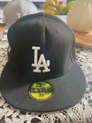 Svart LA Dodgers 59FIFTY keps - Säljer en svart New Era 59FIFTY keps med LA Dodgers-logga i vitt framtill och broderad '1988 World Series' detalj på sidan. Klassisk flat brim och strukturerad passform. Perfekt för dig som gillar sportig streetstyle och vill ha en ikonisk look.